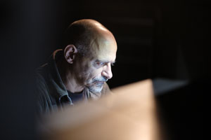 Antoine d’Agata, ici saisi par Pierre Malherbet, dans un moment de concentration intense, plongé dans l’obscurité d’un espace de travail à l’atmosphère feutrée. Éclairé uniquement par une lumière provenant d’un écran ou d’une surface rétroéclairée située face à lui, son visage se détache avec force, révélant les traits marqués du photographe : front dégarni, regard perçant et moustache poivre et sel. Il porte une chemise en velours côtelé sombre sur un t-shirt noir, renforçant l’austérité presque monacale de la scène. L’encadrement resserré et la faible profondeur de champ dirigent toute l’attention sur son regard fixé, absorbé par une tâche probablement numérique ou éditoriale. L’ensemble évoque une immersion totale dans la création, à la croisée de l’intime et de l’acte artistique.